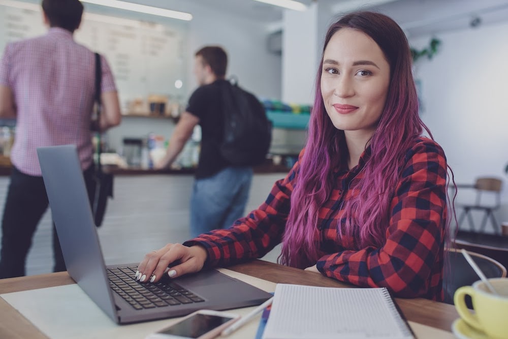 Female college student with purple hair with a laptop and notebook sitting at a table in the cafeteria with students behind her in line getting food.