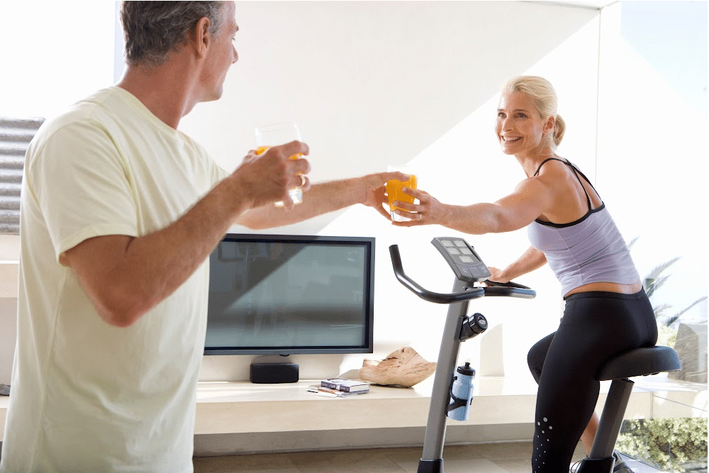 a man over 50 handing a woman over 40 orange juice as she rides an exercise bike.