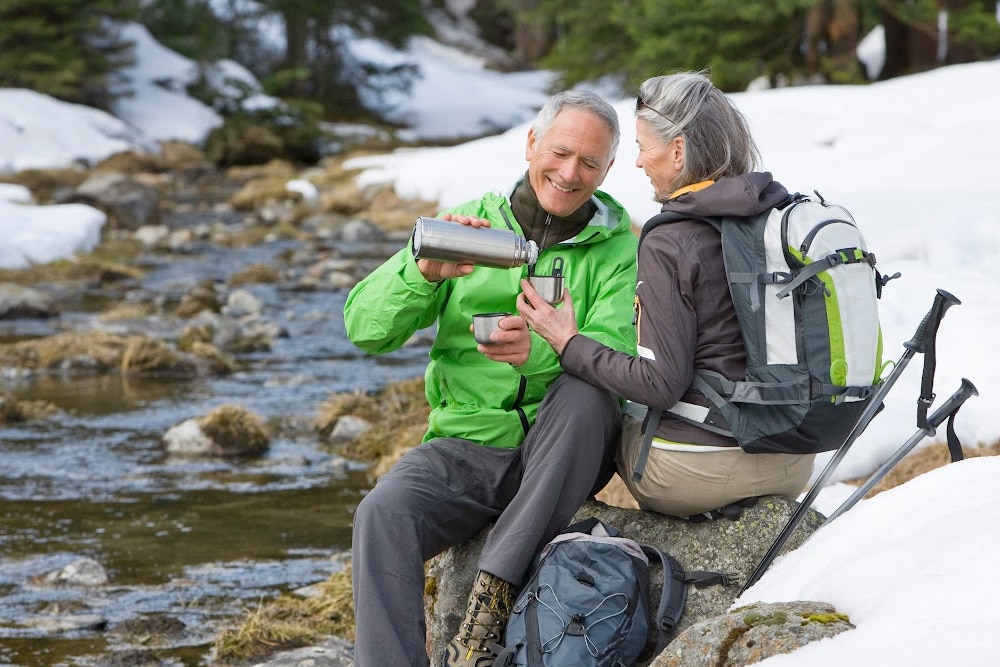 elderly man and a woman over 50 sitting my a creek in the snow drinking hot chocolate taking a break from hiking in the winter with backpacks