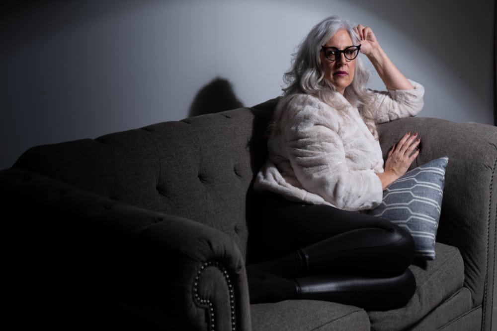 woman over 50 with silver hair and cat eye glasses wearing layers of clothing to keep warm during winter sitting on a sofa