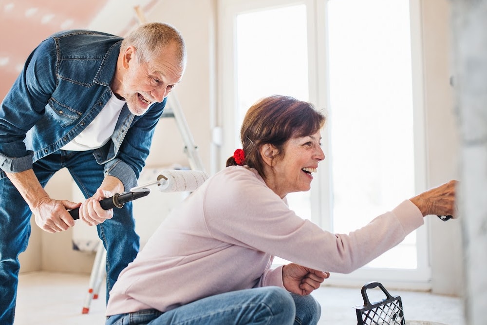 empty nest retired couple painting a room smiling and laughing