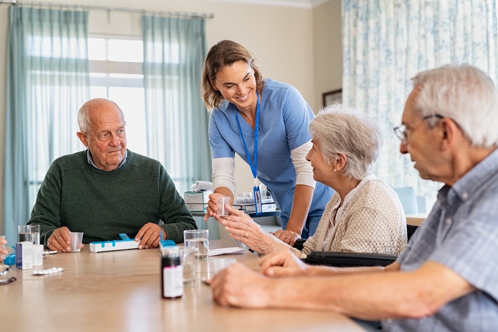 3 residents of assisted living sitting at a table and a nurse is handing them medications.