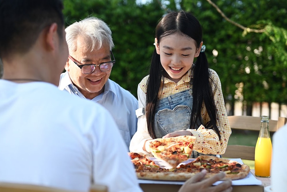 man over 50 with granddaughter and son enjoying pizza outside.