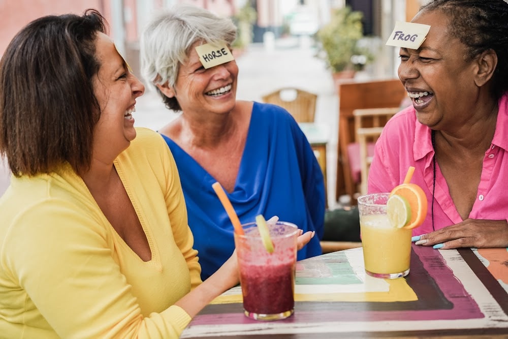 women over 50 having a wonderful summer laughing and playing a game with cards on their foreheads sitting at a table in an outdoor cafe