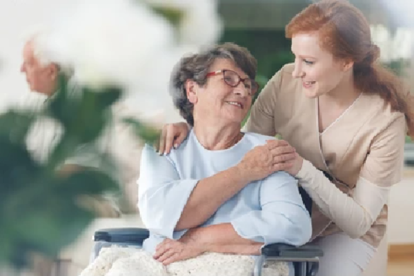 Smiling older woman in a wheelchair holding the hand of a nurse who is leaning over her shoulder smiling.
