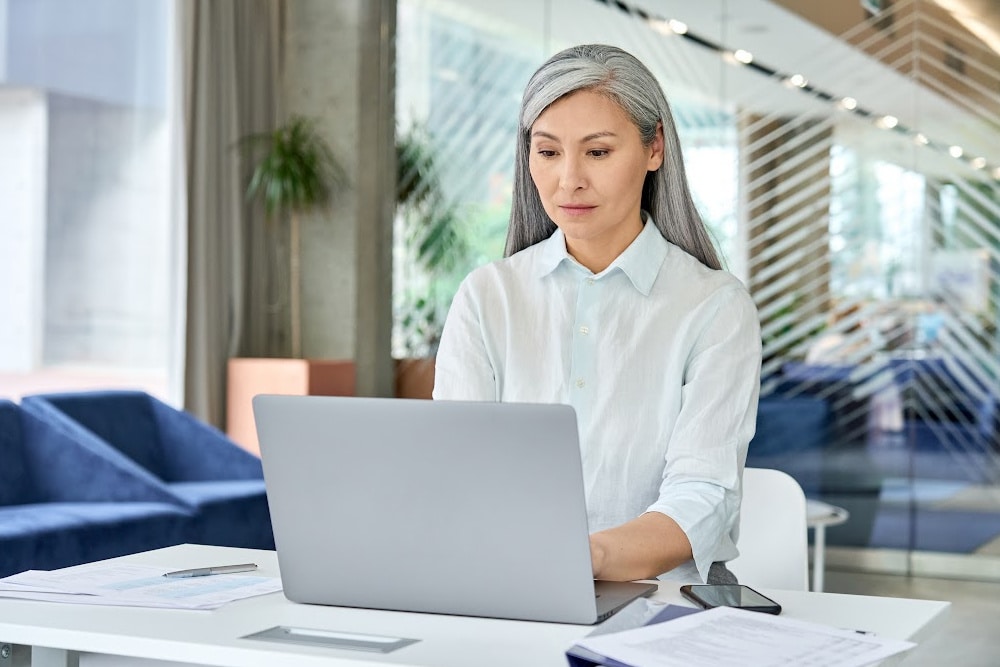 Asian woman over 40 with long silver gray hair setting at a desk in front of a laptop