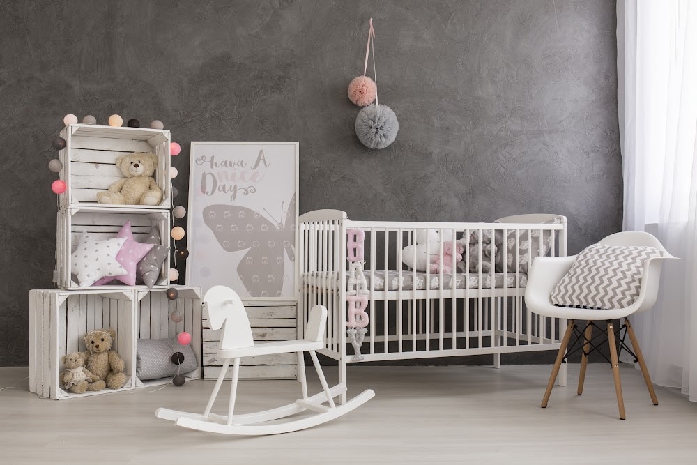 a baby nursery with dark grey textured peel and stick wallpaper and white baby furniture including a bed and a rocking horse. a pink and grey fluffy ball are hanging over the baby bed.