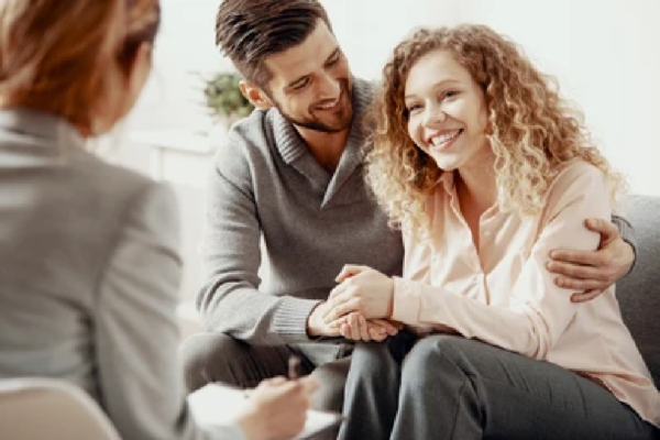 A woman and a man in family therapy. The woman is smiling and the man is holding her had while the therapist looks on supporting her recovery.