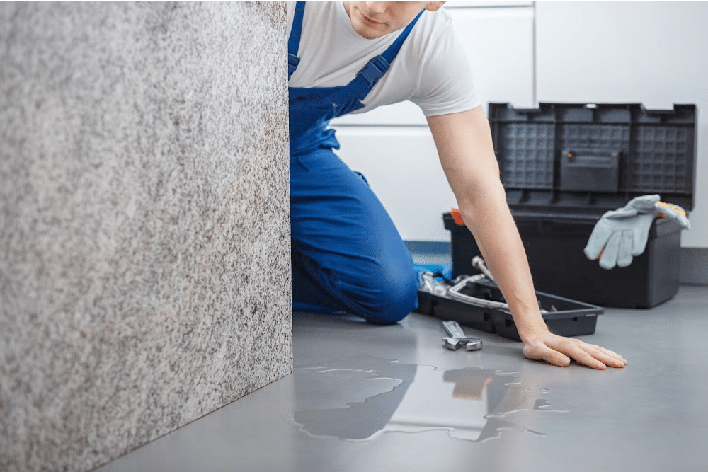 water on the floor from a burst pipe and a person wearing overalls on their knees with a tool box