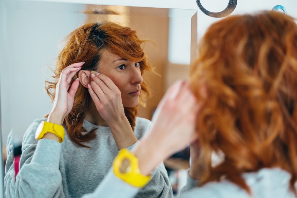 redheaded genx woman wearing a gray sweatshirt and a bright yellow watch putting a hearing aid in her ear while looking in a mirror