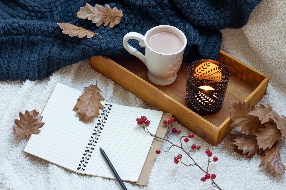 tray with coffee and a lighted candle on a bed beside a notebook and pen and a sweater scattered leaves and berries