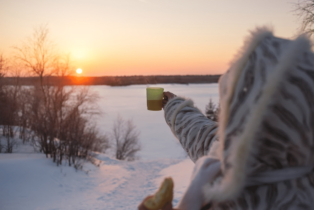 woman over 40 holding a cut of coffee up to the son in a field of snow