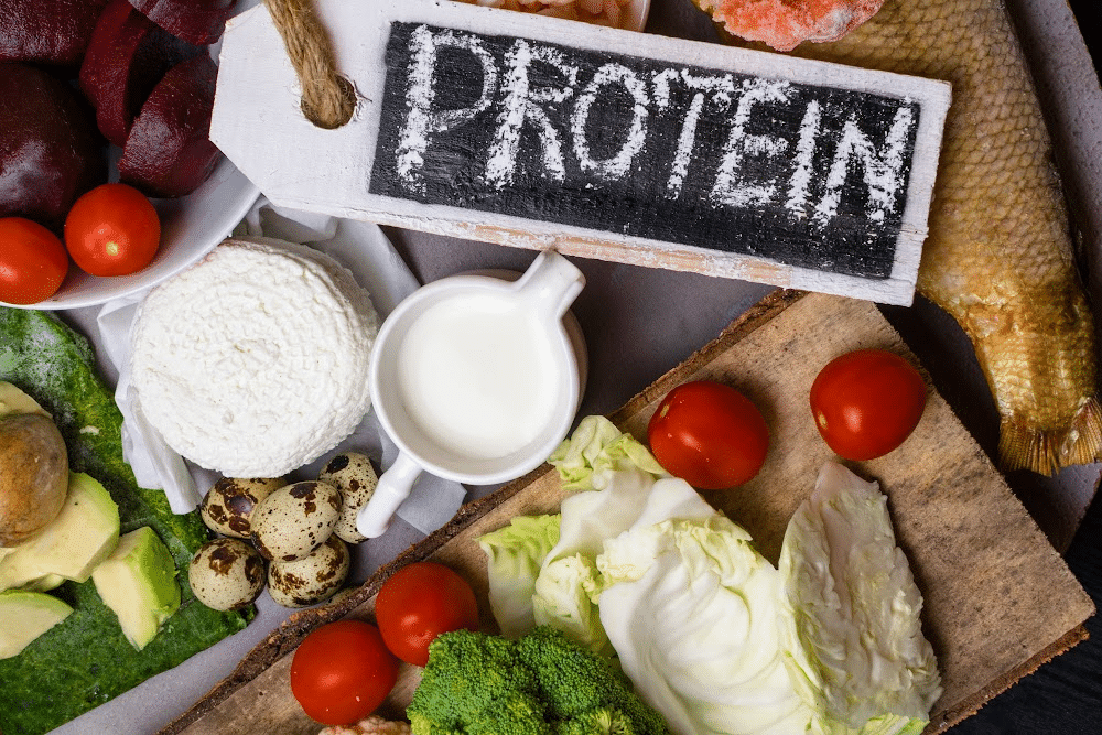 protein written on a chalk board with tomatoes, milk, whey powder, and other foods on a table